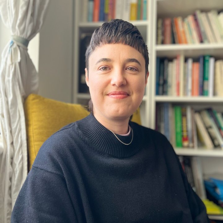 Person sitting indoors with a bookshelf in the background, wearing a black sweater.