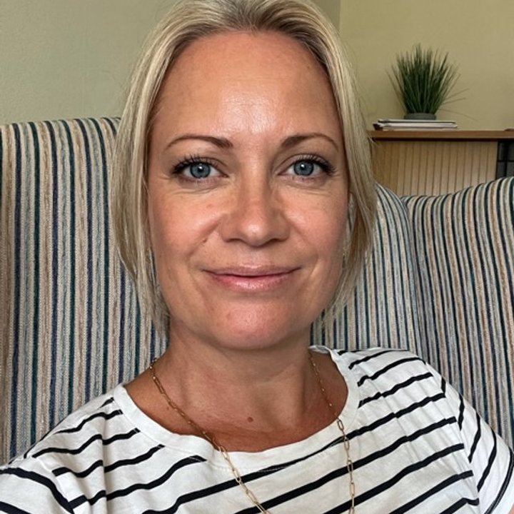 A woman with blonde hair wearing a striped shirt and necklace, sitting on a striped chair with a plant and books in the background.