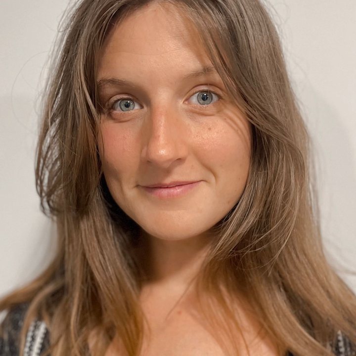 Portrait of a young woman with long brown hair, blue eyes, and a subtle smile, wearing a dark patterned top against a plain white background.
