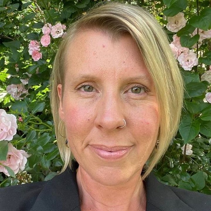 Person in front of pink flowers and green foliage, wearing a black blazer and a gold necklace.
