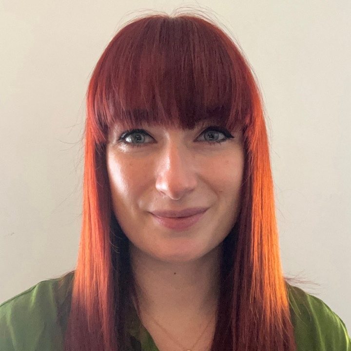 A woman with straight red hair and bangs, wearing a green shirt and a gold necklace with a heart-shaped pendant, smiling at the camera against a plain background.